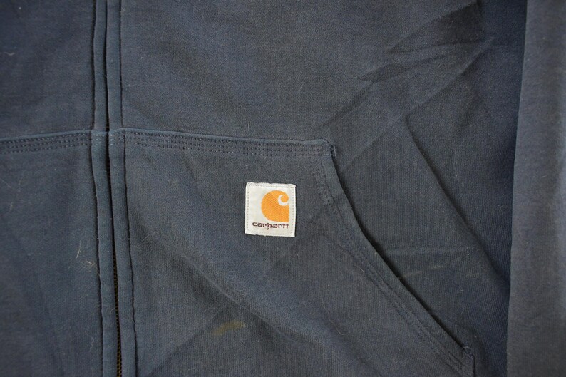 Puede incluir: Primer plano de una sudadera con capucha Carhartt azul marino con cremallera. La sudadera con capucha tiene un bolsillo delantero y una peque&ntilde;a etiqueta cuadrada con el logotipo de Carhartt en naranja y blanco. La cremallera es visible en el centro.