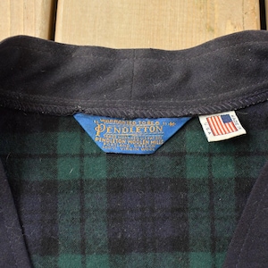 Puede incluir: Primer plano de una bata de lana Pendleton vintage. La bata presenta un cuello azul marino y un estampado de cuadros verdes y azul marino. Se ve una etiqueta azul con letras doradas y una etiqueta con la bandera estadounidense.