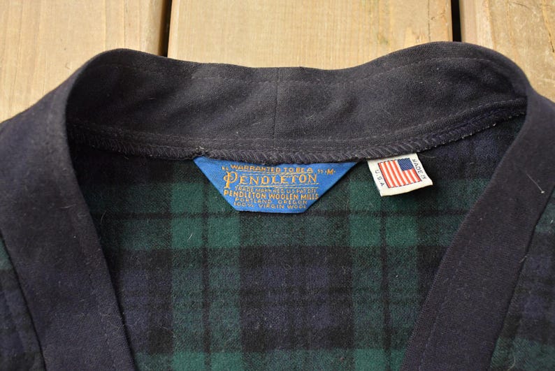 Puede incluir: Primer plano de una bata de lana Pendleton vintage. La bata presenta un cuello azul marino y un estampado de cuadros verdes y azul marino. Se ve una etiqueta azul con letras doradas y una etiqueta con la bandera estadounidense.