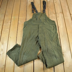 May include: Olive green work overalls with adjustable straps and pockets. The overalls are made of a durable fabric and are laid out on a wooden surface. The bib overalls have a classic design.