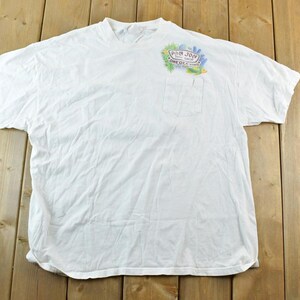 Puede incluir: Camiseta blanca con bolsillo y un gr&aacute;fico colorido en la parte superior izquierda del pecho. El gr&aacute;fico incluye el texto "Pan Jon" y otro texto. La camiseta est&aacute; tendida sobre una superficie de madera.