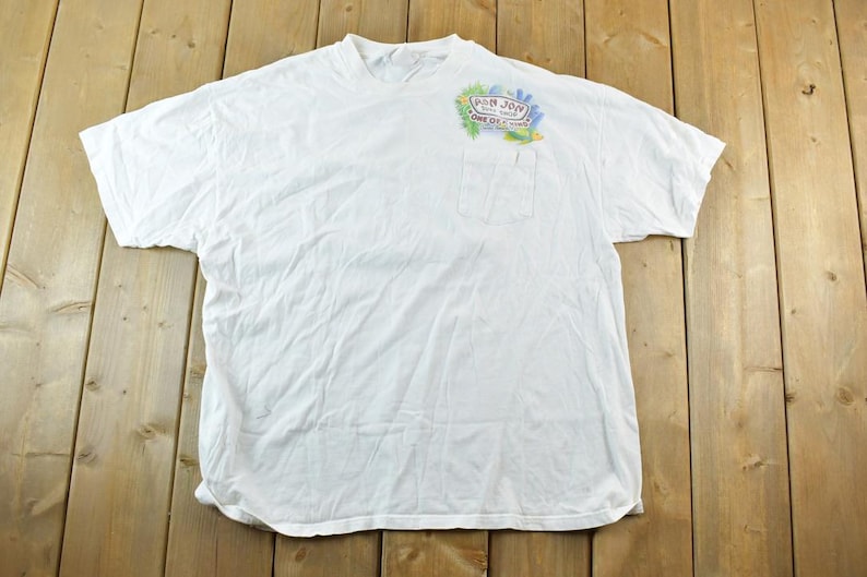 Puede incluir: Camiseta blanca con bolsillo y un gr&aacute;fico colorido en la parte superior izquierda del pecho. El gr&aacute;fico incluye el texto "Pan Jon" y otro texto. La camiseta est&aacute; tendida sobre una superficie de madera.