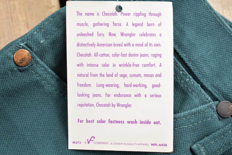May include: A white tag with purple text describes Checotah jeans. The tag says "The name is Checotah. Power rippling through muscle, gathering force. A legend born of unleashed fury. Now, Wrangler celebrates a distinctively American breed with a mind of its own. Checotah. All-cotton, color-fast denim jeans, raging with intense color in wrinkle-free comfort. A natural from the land of sage, sunsets, mesas and freedom. Long-wearing, hard-working, good-looking jeans. For endurance with a serious reputation, Checotah by Wrangler. For best color fastness wash inside out. M275 A COMPANY...A LEADER IN QUALITY APPAREL WPL-6428"