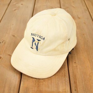 Op de afbeelding: Crèmekleurige Nautica baseballpet met een gebogen klep. De pet heeft de merknaam en het logo in marineblauw geborduurd. De hoed is gemaakt van een zachte, versleten stof en ligt op een houten oppervlak.