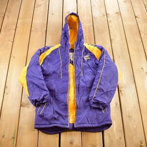 May include: A purple and gold hooded jacket with a zipper closure. The jacket features gold accents on the shoulders, hood, and sleeves. A team logo is visible on the left chest. The jacket is made of a lightweight material.