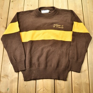 May include: Brown sweater with a yellow stripe across the chest. The sweater has the text "College of St. Francis" embroidered on the front.