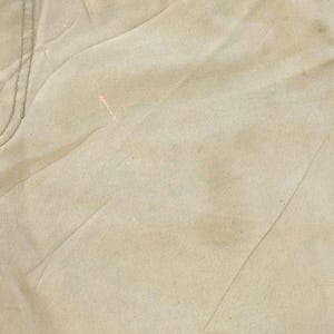 May include: Close-up of tan-colored fabric, possibly denim or canvas, with visible stitching and seams. The fabric has a slightly worn appearance with subtle variations in color and texture, suggesting it has been used.