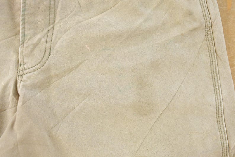 May include: Close-up of tan-colored fabric, possibly denim or canvas, with visible stitching and seams. The fabric has a slightly worn appearance with subtle variations in color and texture, suggesting it has been used.
