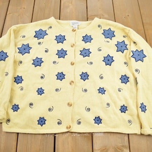 Puede incluir: Un cárdigan amarillo con un patrón de estrellas azules y cierre de botones. El cárdigan tiene un corte holgado y está hecho de una tela suave y cómoda.