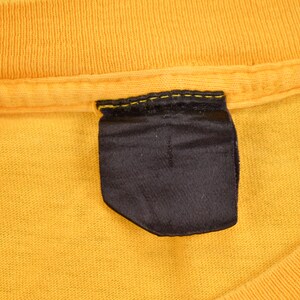 May include: A close-up of a yellow t-shirt with a black fabric label sewn into the neckline.