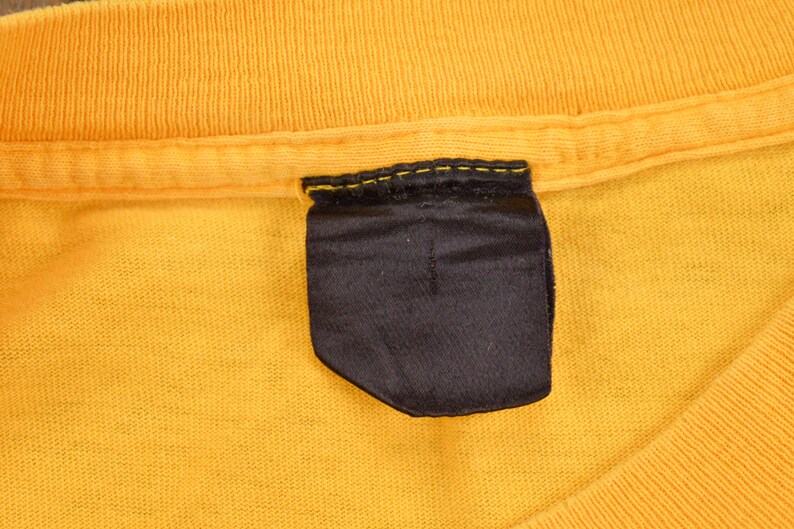 May include: A close-up of a yellow t-shirt with a black fabric label sewn into the neckline.