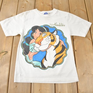 May include: White t-shirt with a colorful graphic of Jasmine from Disney's Aladdin holding Rajah the tiger. The text "Disney Aladdin" is printed on the shirt.