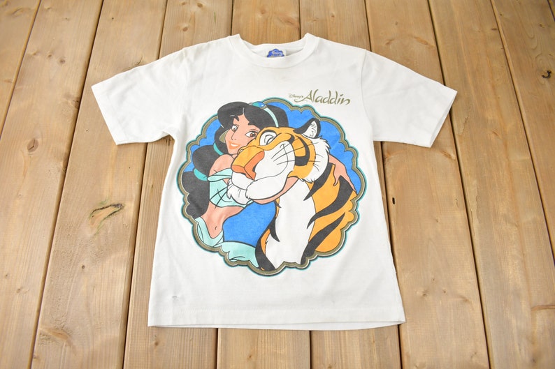 May include: White t-shirt with a colorful graphic of Jasmine from Disney's Aladdin holding Rajah the tiger. The text "Disney Aladdin" is printed on the shirt.