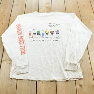 May include: A white long-sleeve t-shirt featuring a graphic of cyclists and the text SIBA 2004 and ARE YOU READY TO RIDE. The left sleeve has the text PUMPKIN PATCH PEDAL printed in orange. The shirt has a crew neckline and ribbed cuffs.
