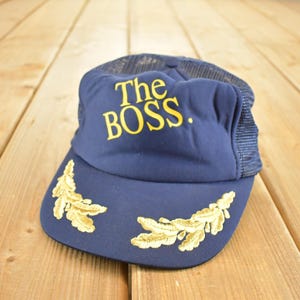 Può includere: Cappellino da camionista blu navy con la scritta "The BOSS" in giallo. Il cappellino ha un retro in rete e disegni di foglie dorate sulla visiera. Un accessorio alla moda.
