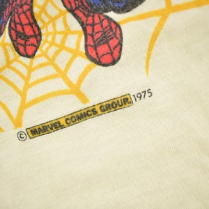 May include: A white t-shirt with a yellow spider web design and a red and blue Spiderman graphic. The shirt has the text "MARVEL COMICS GROUP, 1975" printed on the bottom.