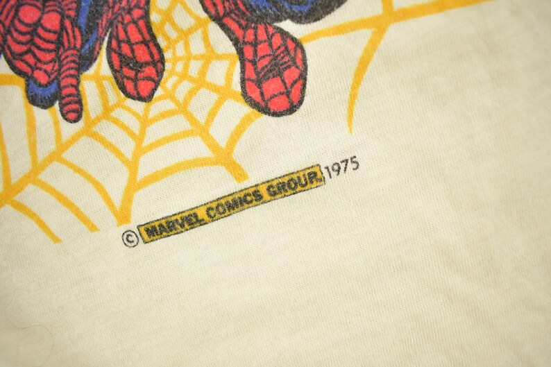 May include: A white t-shirt with a yellow spider web design and a red and blue Spiderman graphic. The shirt has the text "MARVEL COMICS GROUP, 1975" printed on the bottom.