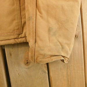 Puede incluir: Primer plano de una chaqueta de trabajo color canela, que muestra el bolsillo, las costuras y la manga. La tela tiene un aspecto desgastado con algunas manchas oscuras. La chaqueta est&aacute; hecha de un material duradero, probablemente lona o una tela similar.