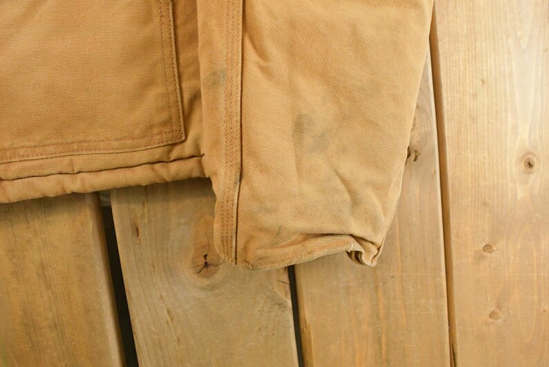 Puede incluir: Primer plano de una chaqueta de trabajo color canela, que muestra el bolsillo, las costuras y la manga. La tela tiene un aspecto desgastado con algunas manchas oscuras. La chaqueta est&aacute; hecha de un material duradero, probablemente lona o una tela similar.