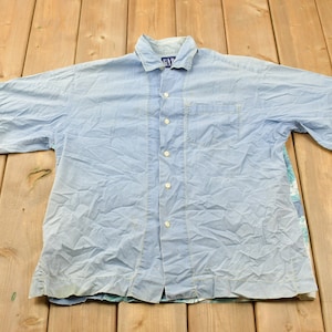 May include: A light blue short-sleeved button-down shirt with a single pocket on the left chest. The shirt is made of a lightweight fabric and has a slightly wrinkled appearance.
