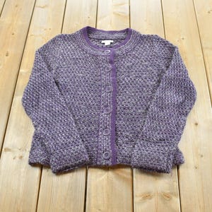 Könnte beinhalten: Eine langärmelige Strickjacke mit Knopfleiste aus einem strukturierten, violett melierten Strickstoff. Das Kleidungsstück verfügt über einen Rundhalsausschnitt, eine einfarbige violette Knopfleiste und passende violette Knöpfe in der Mitte.