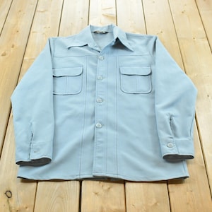 May include: A light blue long-sleeved button-up shirt featuring a pointed collar, two chest pockets with flaps, and vertical stitched detailing running down the front. The garment has matching light blue buttons and buttoned cuffs on each sleeve.