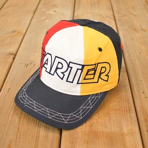 May include: A multi-colored Starter baseball cap. The cap features a navy blue brim and a crown divided into red, white, and yellow panels. The word "STARTER" is prominently displayed in black block letters across the front.