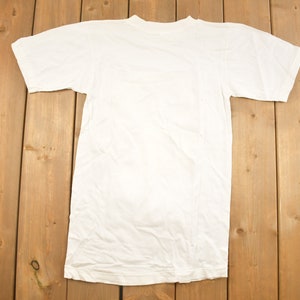 May include: A plain white t-shirt with short sleeves, laid flat on a wooden surface.