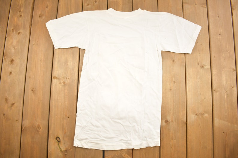 May include: A plain white t-shirt with short sleeves, laid flat on a wooden surface.