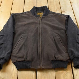 May include: A dark brown and black zip-up bomber jacket featuring a textured fleece-like material, a ribbed collar, ribbed cuffs, and a ribbed hem. The jacket includes two slanted front pockets and a central zipper closure with a visible interior label.