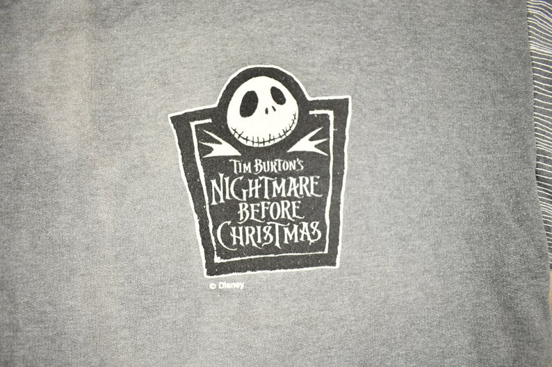 Puede incluir: Camiseta gris con un dise&ntilde;o gr&aacute;fico en blanco y negro de Jack Skellington de "Pesadilla antes de Navidad" de Tim Burton. El dise&ntilde;o incluye el t&iacute;tulo de la pel&iacute;cula. El logotipo de Disney est&aacute; en la parte inferior.