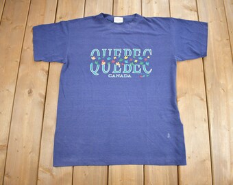 Könnte beinhalten: Ein blaues T-Shirt mit einem weißen Aufdruck, der "Quebec Canada" mit bunten Ahornblättern um den Text herum sagt.