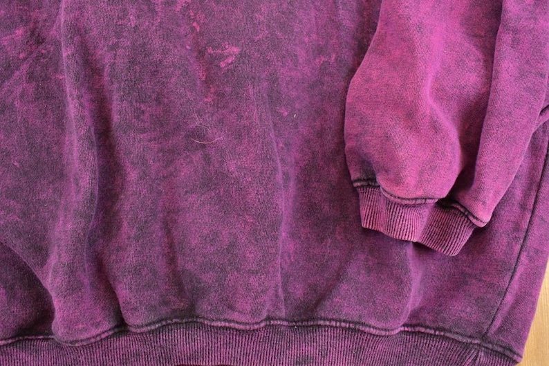 May include: A long-sleeve sweatshirt featuring a deep magenta and black acid-wash pattern. The garment includes a ribbed crew neckline, ribbed cuffs at the end of the sleeves, and a ribbed hemline along the bottom edge, showcasing a textured finish.