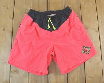 May include: Bright pink shorts with a black waistband and a neon yellow drawstring. The shorts have a black V-shaped panel at the top and a small square patch with the text "Surfs Edge". The shorts are made of a lightweight material.