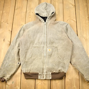 Może przedstawiać: Brązowa kurtka robocza z kapturem, zamkiem błyskawicznym i naszywką z logo Carhartt. Kurtka ma zużyty wygląd z widocznymi śladami użytkowania.