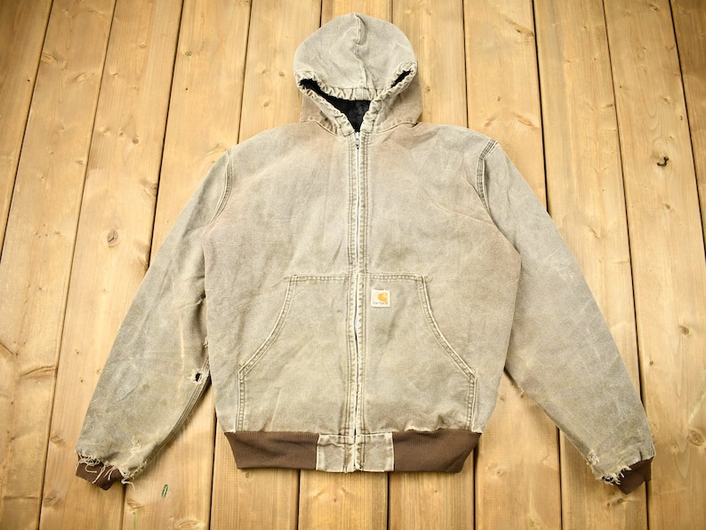 Może przedstawiać: Brązowa kurtka robocza z kapturem, zamkiem błyskawicznym i naszywką z logo Carhartt. Kurtka ma zużyty wygląd z widocznymi śladami użytkowania.