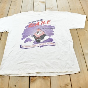 May include: A white short-sleeve t-shirt featuring a graphic of a lion mascot. The text reads Barberton D.A.R.E. St. Augustine and To Resist Drugs, Gangs & Violence. The graphic includes a purple brushstroke background and red and black lettering.