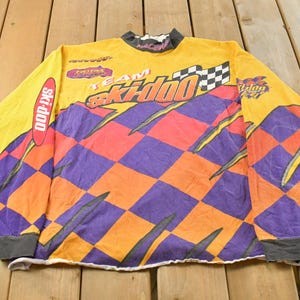 May include: A long-sleeved racing shirt in yellow, orange, and purple, featuring "Team Ski-Doo" graphics and a chequered flag design. The shirt has a high collar and sponsor logos. The sleeves have grey cuffs.