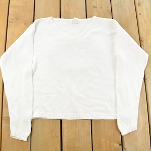 Könnte beinhalten: Ein weißer Pullover mit V-Ausschnitt und langen Ärmeln. Das Strickstück hat gerippte Bündchen und Saum. Der Pullover liegt flach auf einer Holzoberfläche. Hergestellt aus einem weichen Material.