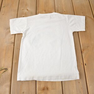 May include: A plain white t-shirt with short sleeves, laid flat on a wooden surface.