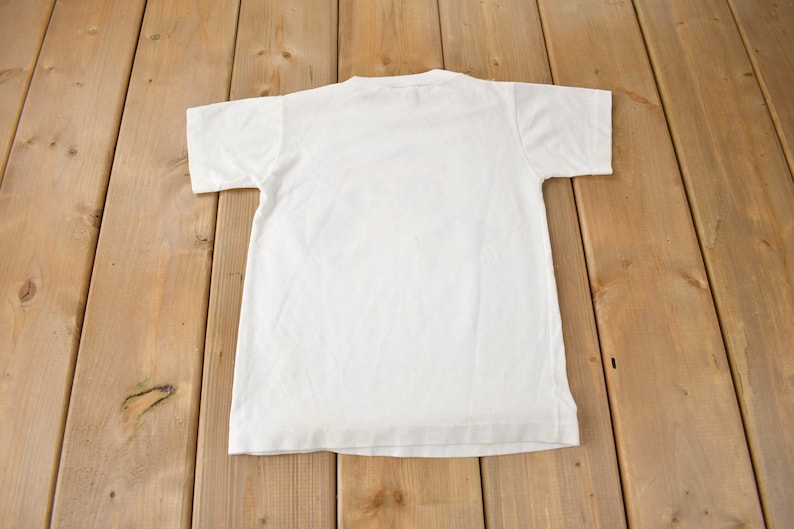 May include: A plain white t-shirt with short sleeves, laid flat on a wooden surface.