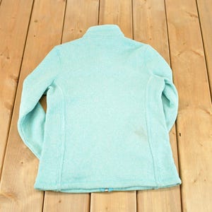 May include: A light blue fleece jacket with a high collar and a full-length zipper. The jacket is made of a soft, textured material and has long sleeves. The back of the jacket is visible, showing the seams and the overall shape.