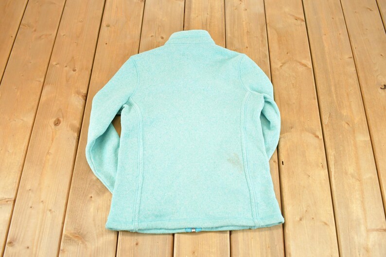 May include: A light blue fleece jacket with a high collar and a full-length zipper. The jacket is made of a soft, textured material and has long sleeves. The back of the jacket is visible, showing the seams and the overall shape.