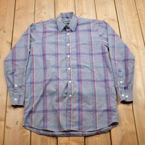 May include: A long-sleeved button-down shirt with a plaid pattern in shades of blue, purple, and white. The shirt has a single chest pocket and a button-down collar.