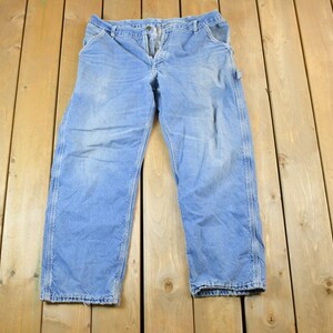 K&ouml;nnte beinhalten: Hellblaue Jeans-Arbeitshose mit Knopfverschluss und mehreren Taschen. Die Hose liegt flach auf einer Holzoberfl&auml;che. Die Jeans hat ein verwaschenes Aussehen und weist Gebrauchsspuren auf.