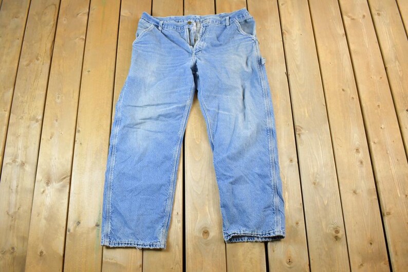 K&ouml;nnte beinhalten: Hellblaue Jeans-Arbeitshose mit Knopfverschluss und mehreren Taschen. Die Hose liegt flach auf einer Holzoberfl&auml;che. Die Jeans hat ein verwaschenes Aussehen und weist Gebrauchsspuren auf.