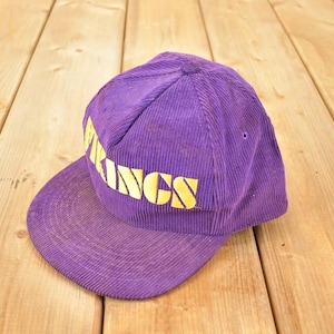 May include: A purple corduroy baseball cap featuring the word "VIKINGS" in yellow lettering. The cap has a flat peak and a structured crown. The hat is on a wooden surface.