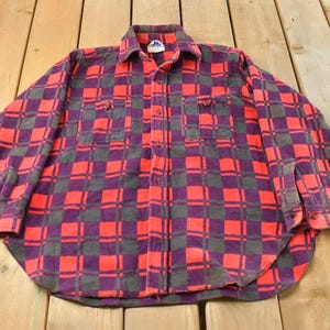 May include: A button-down flannel shirt in a red, purple, and grey plaid pattern. The long-sleeved shirt has two chest pockets and a classic collar. The shirt is laid flat on a wooden surface.