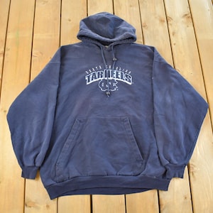 K&ouml;nnte beinhalten: Ein marineblauer Kapuzenpullover mit einer Vordertasche und Kordelzug. Der wei&szlig;e Text auf der Vorderseite lautet "North Carolina Tar Heels" mit einem Logo. Der Pullover liegt auf einer Holzoberfl&auml;che.