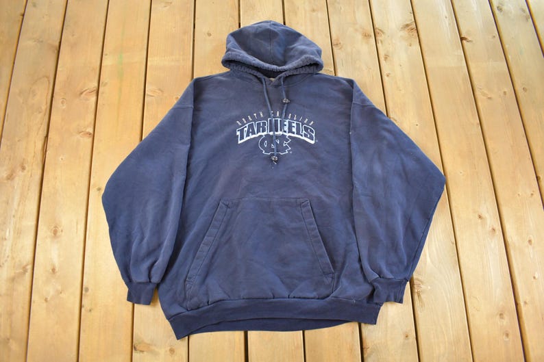 K&ouml;nnte beinhalten: Ein marineblauer Kapuzenpullover mit einer Vordertasche und Kordelzug. Der wei&szlig;e Text auf der Vorderseite lautet "North Carolina Tar Heels" mit einem Logo. Der Pullover liegt auf einer Holzoberfl&auml;che.
